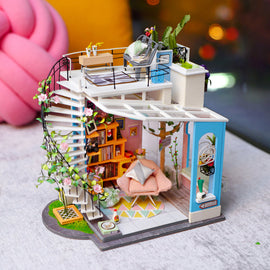 Dora's Loft DIY Model Kit by Robotime is a delightful miniature home model featuring intricate details. It showcases a spiral staircase, cozy living area with bookshelves, a soft pink chair, and vibrant plants. The scene is crafted with artistic touches and realistic furnishings, making it perfect for craft enthusiasts and model builders.