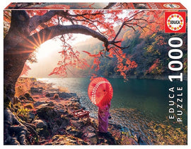 A beautifully illustrated jigsaw puzzle featuring a serene scene of Katsura River in Japan at sunrise. The image depicts a tranquil landscape with vibrant autumn colors of red and orange leaves, a traditional figure holding a red parasol standing by the water's edge, and mountains in the background, all created with 1000 pieces by Educa.