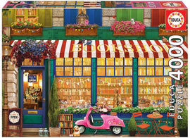A colorful jigsaw puzzle box featuring a charming vintage bookshop. The illustration shows an inviting bookshop facade with red and white striped awnings, vibrant flower boxes, and a pink scooter parked outside. The storefront displays an assortment of books and prints through large windows, while decorative banners add a festive touch.