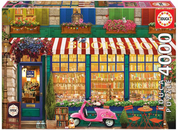 A colorful jigsaw puzzle box featuring a charming vintage bookshop. The illustration shows an inviting bookshop facade with red and white striped awnings, vibrant flower boxes, and a pink scooter parked outside. The storefront displays an assortment of books and prints through large windows, while decorative banners add a festive touch.