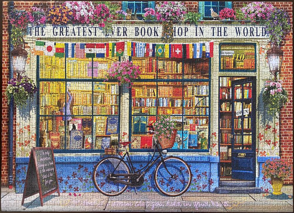 Jigsaw puzzle of a charming bookshop titled 'The Greatest Ever Book Shop in the World.' The image features a flower-adorned facade with colorful flags, a bicycle with a basket, and shelves brimming with books visible through the large windows. The entrance shows a quaint 'Open' sign, inviting visitors into the literary haven.