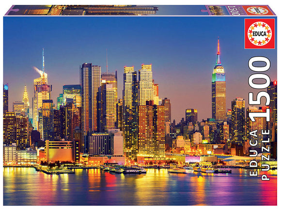 MANHATTAN AT NIGHT 1500PC jigsaw puzzle by Educa, featuring a stunning skyline view of New York City illuminated at night, showcasing iconic buildings like the Empire State Building. The vibrant colors of the city lights reflect off the water, creating a captivating scene for puzzle enthusiasts.