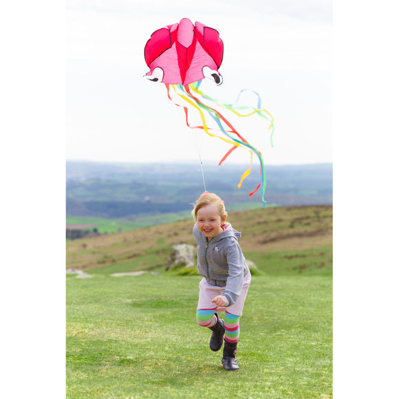 A young girl joyfully running outdoors while flying a vibrant pink octopus kite with colorful ribbons streaming behind it. The setting features rolling green hills under a slightly cloudy sky.