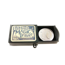 The Wizard's Magic Drawer by House of Marbles features a vintage-style box with intricate lettering and design. The drawer is slightly open, revealing a shiny, round coin marked with '10'. This playful and enchanting item is perfect for magic shows and party tricks.