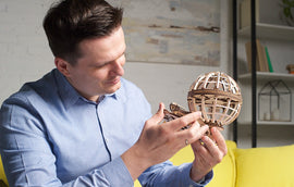 A man is carefully examining the UGears 3D Globe, a beautifully crafted DIY kit made from wood. The intricate details of the globe are highlighted as he holds it in front of him, showcasing its unique design. The scene is set in a bright, modern room with a yellow couch in the background, emphasizing the creativity and craftsmanship of the UGears product.