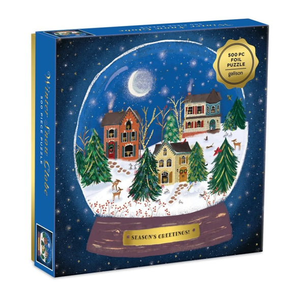 The Winter Snow Globe Foil Puzzle by Galison features a picturesque winter scene within a snow globe. The design showcases cozy houses adorned with holiday decorations, surrounded by snow-covered trees, twinkling stars, and a bright moon. The box displays the text 'Season's Greetings!' and indicates it contains 500 pieces. Ideal for holiday-themed puzzle enthusiasts.
