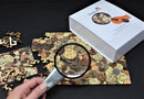 The Bewilderness Coin Maze Wooden jigsaw puzzle is displayed on a dark surface. A hand holds a magnifying glass over a partially completed puzzle featuring intricate coin designs. The box of the puzzle sits beside the puzzle pieces scattered around, illustrating the rich textures and colors of the coins.
