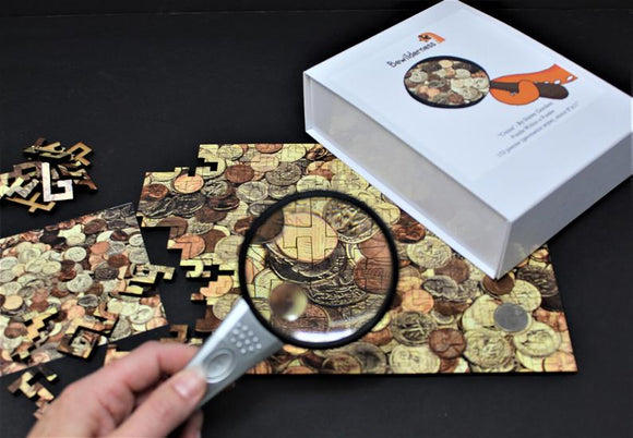 The Bewilderness Coin Maze Wooden jigsaw puzzle is displayed on a dark surface. A hand holds a magnifying glass over a partially completed puzzle featuring intricate coin designs. The box of the puzzle sits beside the puzzle pieces scattered around, illustrating the rich textures and colors of the coins.