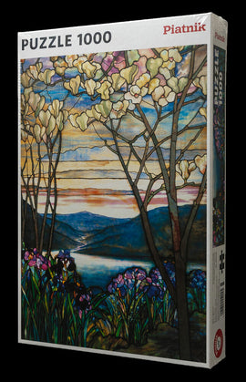 A 1000-piece jigsaw puzzle titled 'Tiffany Magnolia & Irises' by Piatnick displayed in its box. The artwork features vibrant magnolia flowers and irises in a serene landscape, showcasing rich colors and intricate designs. The scene includes trees with blossoms, a flowing river, and distant mountains under a colorful sky at dusk, making it a beautiful and challenging puzzle for enthusiasts.