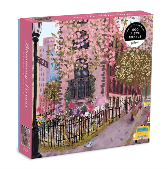 The Blooming Streets jigsaw puzzle by Galison showcases a charming street scene adorned with blossoming trees, vintage buildings, and a welcoming sidewalk lined with blooming flowers. The packaging features a whimsical illustration that invites puzzle enthusiasts to immerse themselves in a picturesque urban landscape. This 500-piece puzzle brings together art and the joy of puzzling, offering an entertaining activity for all ages.