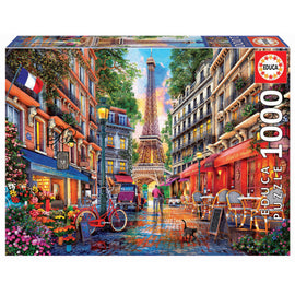 Educa jigsaw puzzle titled 'PARIS; DOMINIC DAVISON'. The image features a vibrant Parisian street scene with the Eiffel Tower in the background, bustling cafés, colorful buildings, a red bicycle, and a cat, all set against a sunset sky.