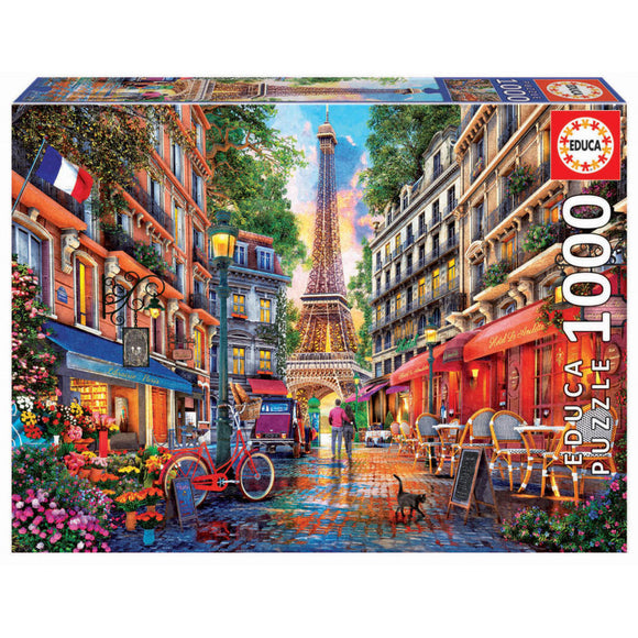 Educa jigsaw puzzle titled 'PARIS; DOMINIC DAVISON'. The image features a vibrant Parisian street scene with the Eiffel Tower in the background, bustling cafés, colorful buildings, a red bicycle, and a cat, all set against a sunset sky.