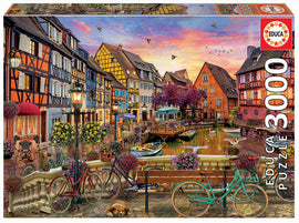 EDUCA 3000-piece jigsaw puzzle featuring a picturesque scene of Colmar, France. The artwork showcases colorful timber-framed houses, lush flowered balconies, bicycles parked by the canal, and reflections in the water at sunset. Includes charming details like a dog resting and birds flying above, providing a delightful representation of a serene French village.