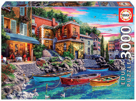 A vibrant 3000-piece jigsaw puzzle titled 'Sunset in Como' by Educa. The image features a picturesque lakeside scene with colorful boats docked near charming buildings, surrounded by lush greenery and a clear blue sky. The puzzle artwork captures the serene beauty of a sunset reflecting on the water, inviting puzzle enthusiasts to enjoy a relaxing challenge.