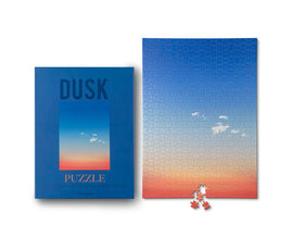 The Wonders of Nature Dusk jigsaw puzzle by Printworks features a stunning gradient image of a sunset in soothing blue and orange tones. The puzzle comes in a beautifully designed box that showcases a portion of the puzzle image, alongside some scattered puzzle pieces, ready for assembly. This puzzle promises a calming and engaging experience for enthusiasts of all ages.