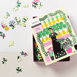 Seltzer Goods' Fruit Lady jigsaw puzzle featuring a colorful illustration of a woman surrounded by fruits, plants, and vibrant patterns. The 500-piece puzzle is displayed partially completed, with a box showing the cover design alongside scattered puzzle pieces.