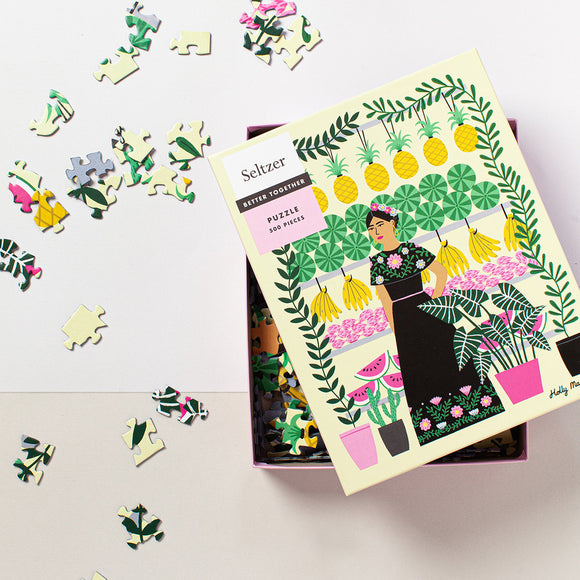 Seltzer Goods' Fruit Lady jigsaw puzzle featuring a colorful illustration of a woman surrounded by fruits, plants, and vibrant patterns. The 500-piece puzzle is displayed partially completed, with a box showing the cover design alongside scattered puzzle pieces.