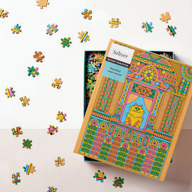 A vibrant jigsaw puzzle box featuring a colorful design with intricate patterns and a central image of a stylized house cat. The box is opened to reveal scattered puzzle pieces around it, showcasing various colors and shapes. The packaging is from Seltzer Goods and indicates a 1000-piece puzzle. The background is a light, neutral color which emphasizes the bright artwork on the box.