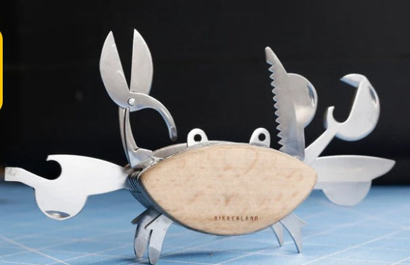 A whimsical Crab Multi Tool by Kikkerland with various stainless steel tools integrated into a crab design, including scissors, a screwdriver, and other utility functions, all set against a simple dark background. The body's surface has a natural wood finish, adding a unique touch.