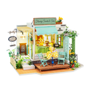 A detailed model kit of a quaint tea and sweets shop, showcasing vibrant yellow and green accents. The shop features outdoor seating with two turquoise chairs and a yellow checkered table. Intricate designs include flower decorations and a welcoming sign that reads 'Flawery Sweets & Teas,' creating a charming ambiance perfect for tea lovers and DIY enthusiasts.