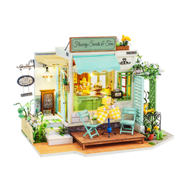 A detailed model kit of a quaint tea and sweets shop, showcasing vibrant yellow and green accents. The shop features outdoor seating with two turquoise chairs and a yellow checkered table. Intricate designs include flower decorations and a welcoming sign that reads 'Flawery Sweets & Teas,' creating a charming ambiance perfect for tea lovers and DIY enthusiasts.