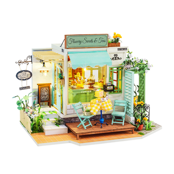 A detailed model kit of a quaint tea and sweets shop, showcasing vibrant yellow and green accents. The shop features outdoor seating with two turquoise chairs and a yellow checkered table. Intricate designs include flower decorations and a welcoming sign that reads 'Flawery Sweets & Teas,' creating a charming ambiance perfect for tea lovers and DIY enthusiasts.