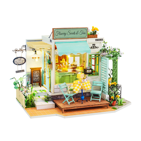 A detailed model kit of a quaint tea and sweets shop, showcasing vibrant yellow and green accents. The shop features outdoor seating with two turquoise chairs and a yellow checkered table. Intricate designs include flower decorations and a welcoming sign that reads 'Flawery Sweets & Teas,' creating a charming ambiance perfect for tea lovers and DIY enthusiasts.
