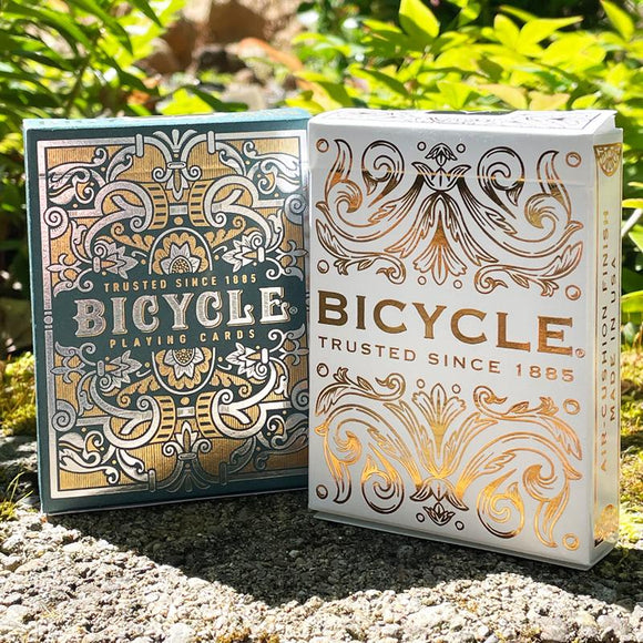 A beautifully designed Bicycle playing card deck featuring two packs: one with an ornate green and gold design and the other in white with elegant gold detailing. The cards are presented outdoors, surrounded by greenery, emphasizing their classic appeal and high quality. Both packs display the 'Trusted Since 1885' slogan, highlighting the brand's long-standing reputation.