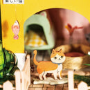 A colorful DIY miniature cat house by Robotime, featuring a charming outdoor scene with a paper cat standing in front of a bright yellow archway. The background includes a cozy sleeping nook, a woven basket, and decorative plants, creating a whimsical environment for cat lovers.