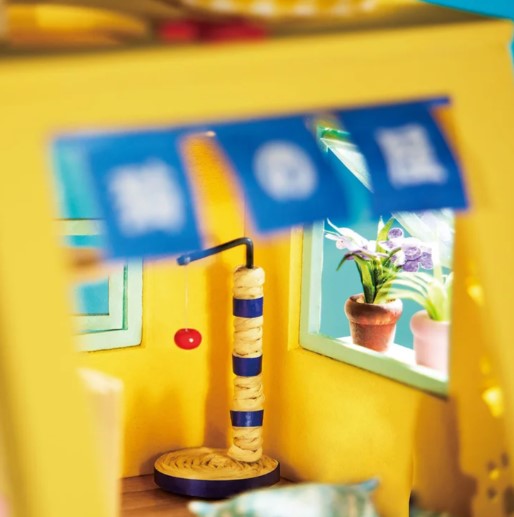 A brightly colored miniature cat house from Robotime, showcasing an engaging interior view with a colorful scratching post, a hanging toy, and window plants. This DIY kit features intricate details, making it a perfect project for cat lovers and miniature enthusiasts.