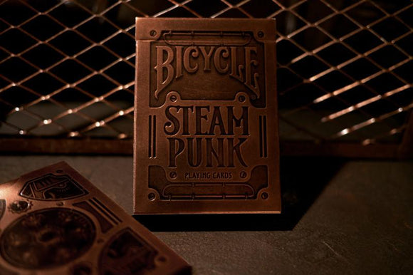 A close-up of the Bicycle Steam Punk Bronze Deck playing cards by Theory 11. The bronze-colored card box features intricate steampunk design elements with the words 'Bicycle STEAM PUNK' prominently displayed. In the background, a textured surface adds depth to the image, emphasizing the vintage aesthetic of the deck.