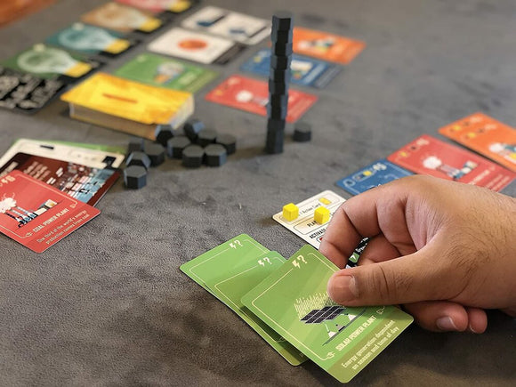 A close-up of a person's hand holding game cards for 'Power Failure' by ARTANA GAMES. The table is covered with various colorful cards, tokens, and a small tower made of hexagonal pieces, showcasing the multi-faceted gameplay.