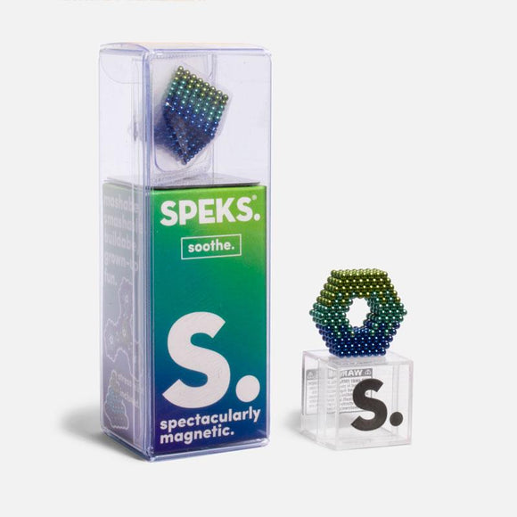 A transparent packaging of Speks Soothe Gradient featuring a colorful, gradient magnetic fidget toy. The display includes a solid base with a hexagonal shape made of magnetic beads in shades of blue and green. The box has branding that indicates the product is 'spectacularly magnetic' with a playful design, suggesting endless building possibilities for stress relief and creativity.