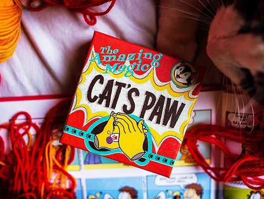 A vibrant box titled 'Cat's Paw' by Ellusionist, designed for magic tricks. The box features colorful graphics and illustrations related to magic, surrounded by red and orange yarns, indicative of a fun creative environment. Part of a playful scene with hints of other items related to magic and performance.