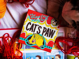A vibrant box titled 'Cat's Paw' by Ellusionist, designed for magic tricks. The box features colorful graphics and illustrations related to magic, surrounded by red and orange yarns, indicative of a fun creative environment. Part of a playful scene with hints of other items related to magic and performance.