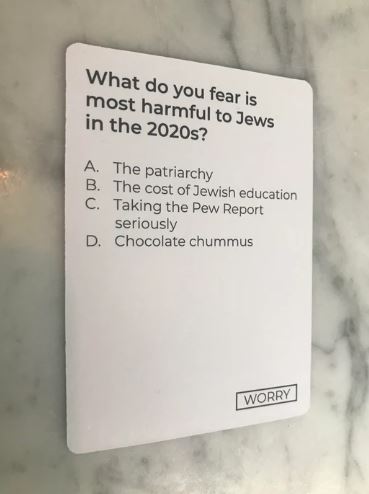 A game card from the Jewish Card Revoked game by Flying Leap Games. The card poses a humorous question about concerns facing the Jewish community in the 2020s, with four multiple-choice options including: 'The patriarchy,' 'The cost of Jewish education,' 'Taking the Pew Report seriously,' and 'Chocolate hummus.' The card is designed to provoke thought and laughter in a group setting.