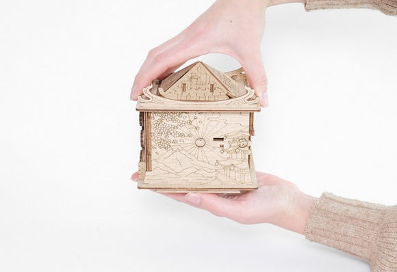 A person holds a detailed mechanical puzzle shaped like a small house, crafted from wood. The intricate designs include landscapes and celestial elements, showcasing a blend of creativity and engineering.