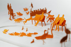 A set of intricately crafted orange paper figures depicting a zoo scene. Various animal shapes like cats, birds, and a group of people sitting on benches, creating a playful and artistic representation of a zoo environment.