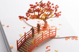 A beautifully crafted 3D model depicting a serene fall scene. Two figures walk across a red wooden bridge surrounded by vibrant autumn leaves and a sculpted tree with colored foliage. The intricate details capture the essence of fall foliage in a stunning DIY kit by TERADA MOKEI.