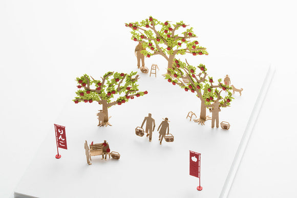 The Apple Picking Model by TERADA MOKEI features intricately designed paper trees laden with red apples. Two human figures are depicted walking with baskets, set against a backdrop of a bench and decorative signs, creating a charming autumn scene. Perfect for DIY enthusiasts, this model brings a touch of whimsy to any space.