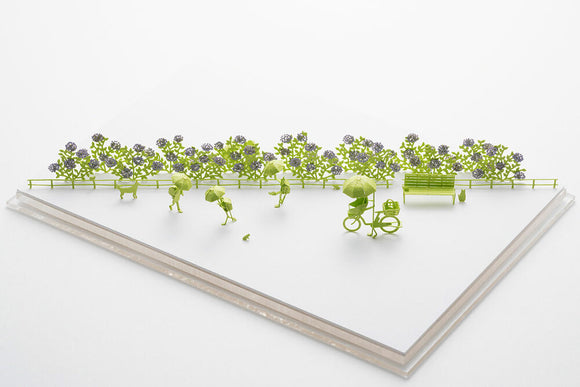 A vibrant and intricate model titled 'Rainy Season' by TERADA MOKEI, featuring miniature figures with umbrellas, a bicycle, and colorful flowers. The scene captures a park-like setting, perfect for DIY enthusiasts who enjoy crafting unique displays.