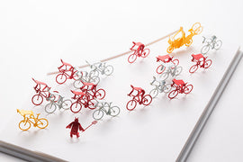 A colorful display of miniature bicycles, featuring various designs in red, yellow, and gray, along with a small figurine in red resembling a devil character. The bicycles are arranged playfully on a white surface, showcasing their intricate details and vibrant colors, perfect for hobbyists and DIY enthusiasts.