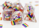 A vibrant Rainbow Flag RPG Dice Set by Foam Brain featuring colorful dice filled with rainbow stripes and white numeric markings. The set includes various polyhedral shapes laid out against a plain background with small rainbow flags scattered around.