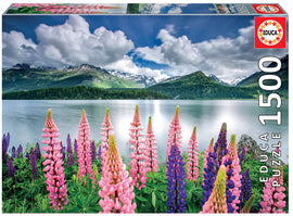 Educa Jigsaw Puzzle featuring a serene lake landscape with vibrant pink and purple flowers in the foreground, snowy mountains in the background, and a cloudy sky reflecting on the still water. This 1500 piece puzzle offers an immersive challenge for puzzle enthusiasts.