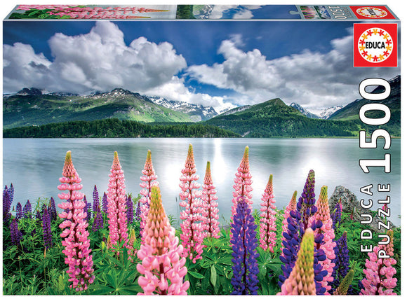 Educa Jigsaw Puzzle featuring a serene lake landscape with vibrant pink and purple flowers in the foreground, snowy mountains in the background, and a cloudy sky reflecting on the still water. This 1500 piece puzzle offers an immersive challenge for puzzle enthusiasts.