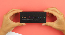 A person holding the FlipBokui: Molecularis DIY kit from Boku Flipbooks. The kit features a sleek black box with colorful lettering 'Molecularis' and a subtitle that reads 'An animated challenge colored by you'. The background is a vibrant pink, enhancing the aesthetic appeal of the product.
