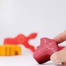 A person holding a vibrant red wooden puzzle piece with floral engravings, part of the Casa Glora collection by Grimm's. In the background, blurred colorful pieces are visible, highlighting the playful nature of the jigsaw puzzle. The piece displays craftsmanship and encourages creative play.