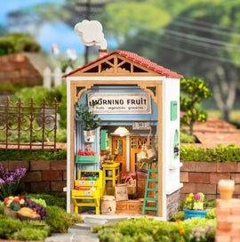 A detailed model kit of a small fruit store named 'Morning Fruit'. The building features a colorful facade with a sign displaying 'Morning Fruit', shelves filled with fruits and vegetables, and a welcoming atmosphere. The exterior has vibrant colors, potted plants, and a ladder, creating a charming scene in a garden setting.