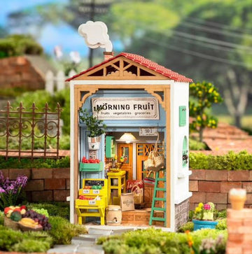 A detailed model kit of a small fruit store named 'Morning Fruit'. The building features a colorful facade with a sign displaying 'Morning Fruit', shelves filled with fruits and vegetables, and a welcoming atmosphere. The exterior has vibrant colors, potted plants, and a ladder, creating a charming scene in a garden setting.