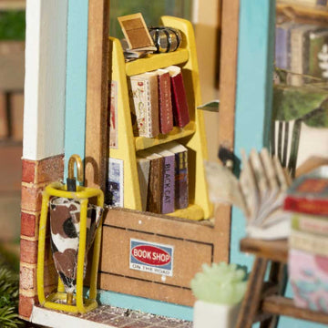A detailed view of the Free Time Bookshop model kit by Robotime, showcasing a miniature yellow bookshelf filled with tiny books and a decorative shop front sign that reads 'BOOK SHOP ON THE ROAD'. The scene features a charming and cozy setup that captures the essence of a quaint bookshop, complete with a small plant and a unique decorative element in the foreground.